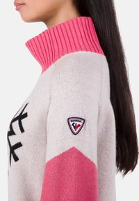 Sweter robiony na drutach w jasnoszarym kolorze z różowym ściągaczem, czarnym wzorem oraz naszywką z logo Rossignol na lewym rękawie.