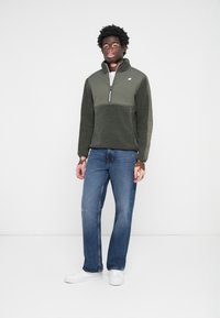 Green fleece pullover with a zipper and a contrasting upper panel, paired with blue jeans and white sneakers, set against a white background.