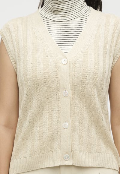 Μπεζ αμάνικη πλεκτή γιλέκο με κουμπιά, φορεμένη πάνω από μαύρο και άσπρο ριγέ ζιβάγκο.