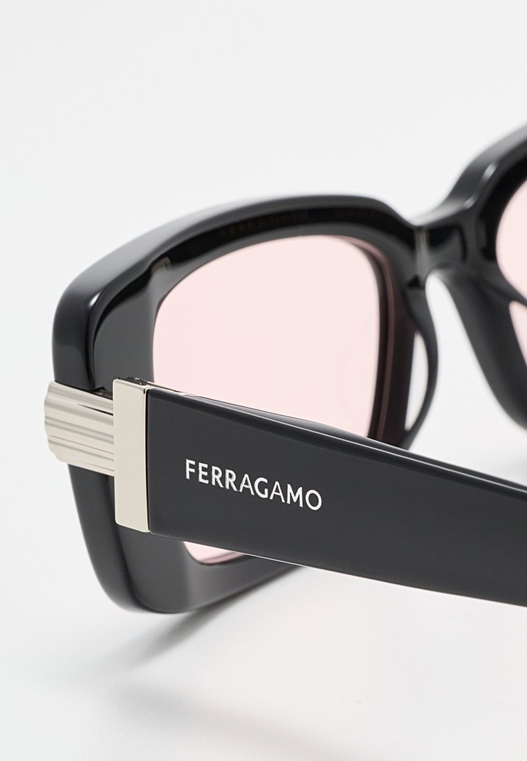 Gafas de sol negras de gran tamaño con lentes rosas. Presentan un marco brillante y un detalle de logo plateado en las patillas. Forma rectangular.