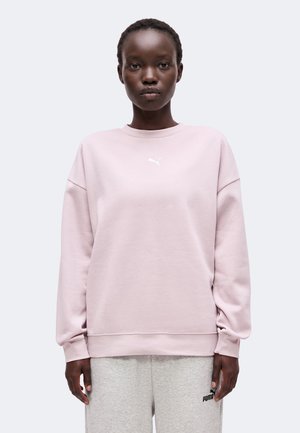 Persona que lleva una sudadera rosa claro de Puma y pantalones deportivos grises claros de Puma, de pie mirando hacia adelante sobre un fondo liso.