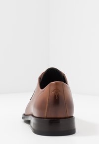 Chaussure habillée en cuir brun avec une finition lisse, bout arrondi et logo en relief sur le talon. Semelle en caoutchouc noire légèrement brillante.