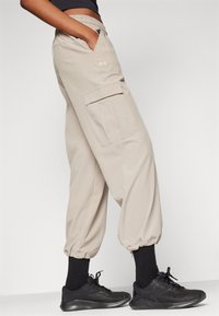 Pantaloni cargo beige con polsini ed elastico in vita. Presentano una tasca laterale e un tessuto morbido, abbinati a scarpe athletic nere e calzini.