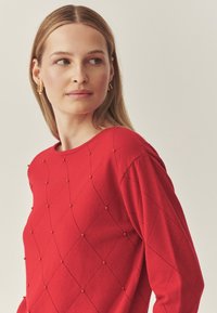 Piros kötött pulóver gyémántmintával, apró díszítésekkel, kerek nyakkivágással és hosszú ujjakkal. Puha textúrájú és testhezálló fazon.