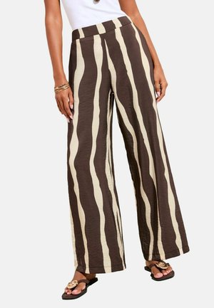 Sieviete, kas valkā plaša piegriezuma bikses ar augstu vidukli un neregulārām vertikālām brūnām un bēšām svītrām, saskaņotas ar brūnām sandales un baltu topu.