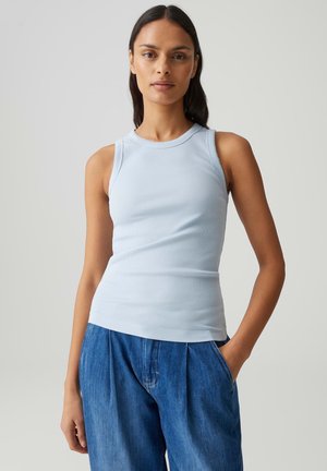 Vrouw die een lichtblauw, mouwloos geribd shirt en losse blauwe spijkerbroek draagt, staand met één hand in haar zak tegen een effen achtergrond.