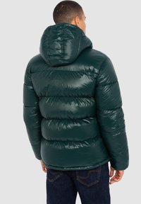 Chaqueta inflable verde con acabado brillante, con costuras horizontales y capucha, llevada sobre unos vaqueros azul oscuro. Puños ajustables.