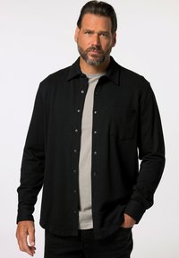 Camisa negra de manga larga con botones, hecha de una tela suave, que presenta un bolsillo en el pecho, cuello clásico y botones plateados. Lleva puesta una camisa gris debajo.