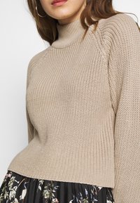 Beżowy sweter z wysokim kołnierzem i prążkowaną fakturą, o luźnym kroju. W zestawie z czarną spódnicą w kwiatowy wzór.