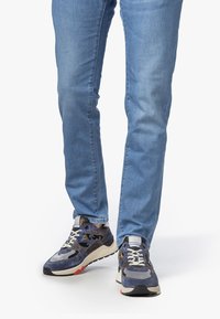 Henkilö, jolla on yllä siniset farkut ja tummansiniset tennarit vaaleilla pohjilla ja valkoisilla nauhoilla, seisoo valkoista taustaa vasten.