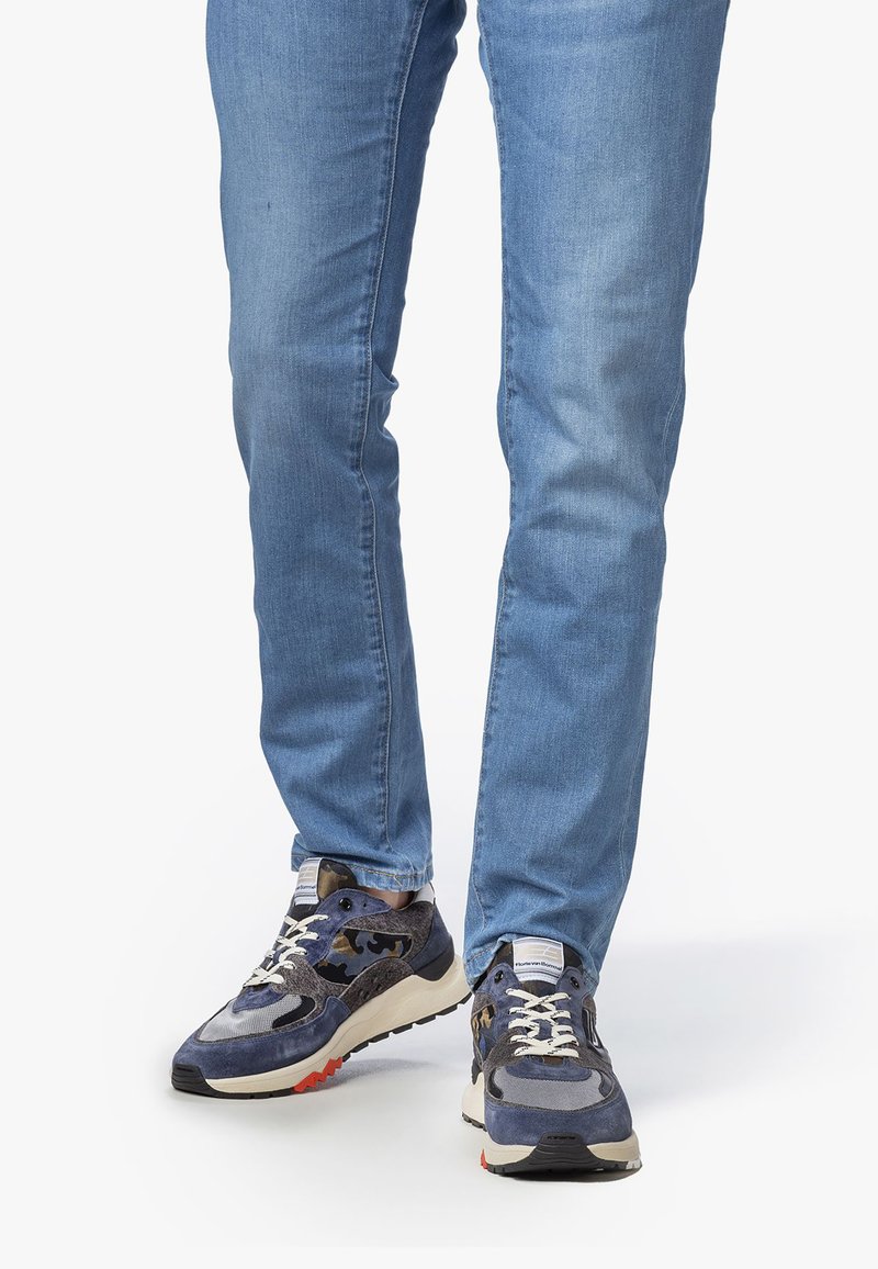 Henkilö, jolla on yllä siniset farkut ja tummansiniset tennarit vaaleilla pohjilla ja valkoisilla nauhoilla, seisoo valkoista taustaa vasten.