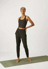 Nő mezítláb áll egy zöld jogamatracon, fekete ujjatlan felsőt és fekete sportnadrágot visel, az egyik kezét a csípőjén tartja.