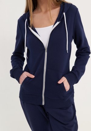 Femme portant un sweat à capuche zippé bleu marine avec cordons blancs, les mains dans les poches, assorti d'un pantalon bleu marine et d'un haut blanc.