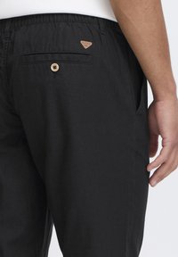 Zwarte broek met een relaxte pasvorm, gemaakt van lichtgewicht stof. Bevat een achterzak met een houten knop en een leren patch op de taille.