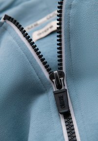 Jasnoniebieska tkanina z teksturowanym splotem, z czarnym zapięciem na zamek oraz białymi przeszyciami wzdłuż krawędzi. Wewnątrz widoczna metka.