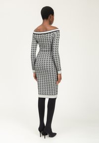 Frau mit dem Rücken zu sehen, in einem taillierten, schulterfreien schwarz-weißen gemusterten knielangen Kleid, schwarzen Strumpfhosen und schwarzen High Heels.