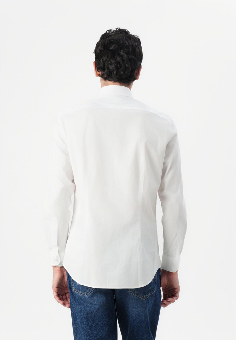 Chemise blanche à manches longues avec un col standard, texture lisse et ourlet arrondi, portée avec un jean bleu. Pas de motifs ou de logos visibles.