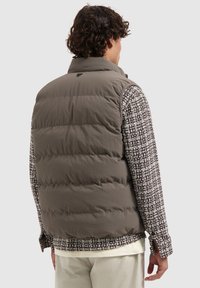 Braune Steppweste mit hohem Kragen und gestepptem Design. Getragen über einem strukturierten braun-weißen gemusterten Hemd.
