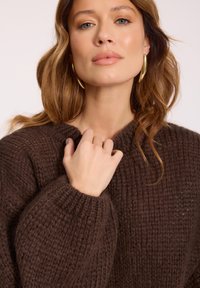 Brązowy sweter dziany z teksturowanym wzorem, luźny krój i szerokie rękawy. Projekt w serek, z ręką na obrzeżu kołnierza.