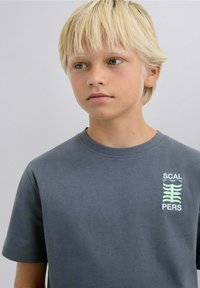 Camiseta de algodón gris con mangas cortas, que presenta un diseño impreso en texto verde y blanco en el lado izquierdo del pecho.