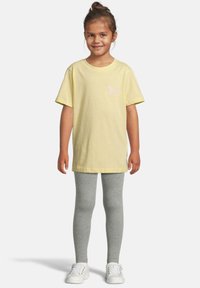 Jeune fille souriante debout, portant un t-shirt jaune avec un papillon, un legging gris et des baskets blanches sur un fond uni.