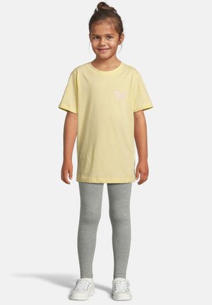 Smiling young girl standing, wearing a yellow t-shirt with a butterfly, gray leggings, and white sneakers against a plain background.