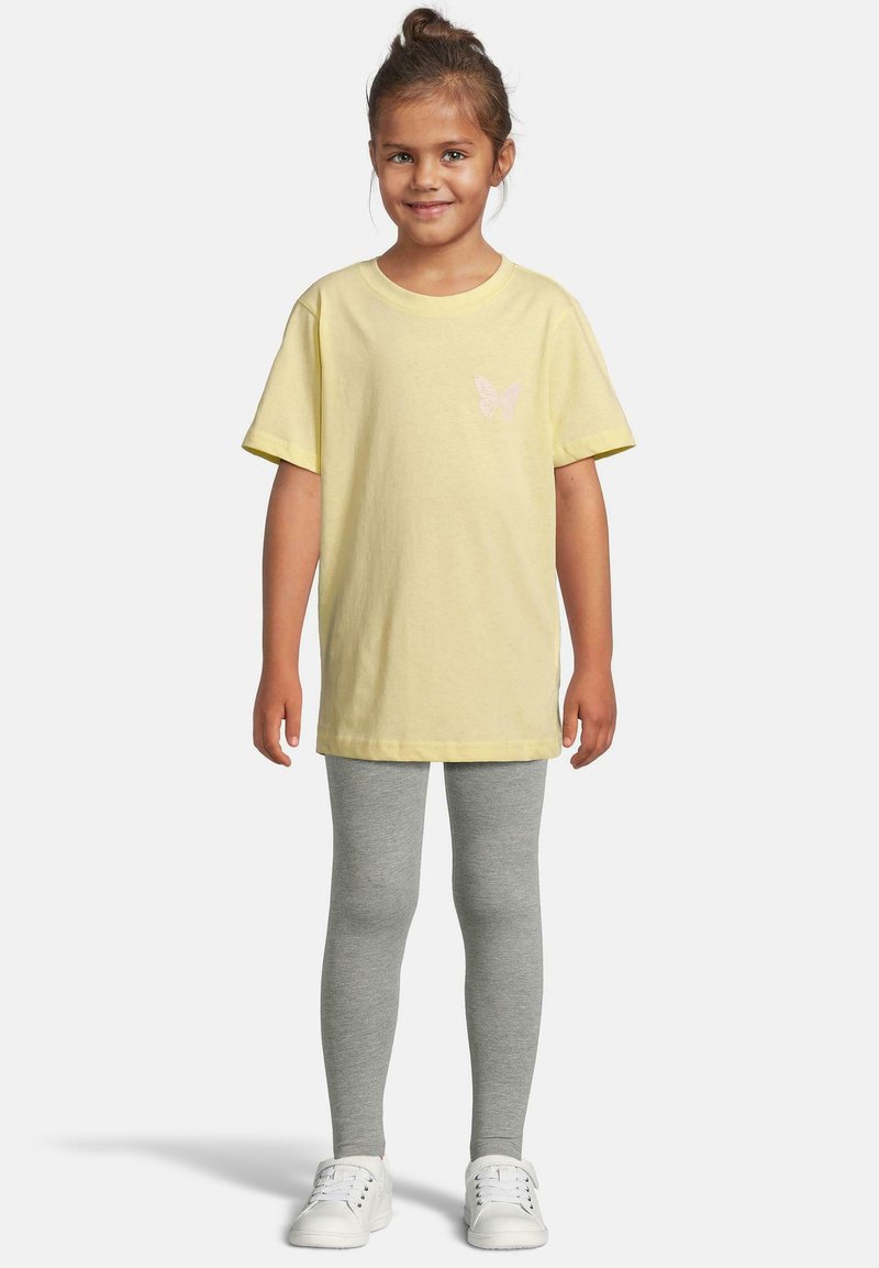 Jeune fille souriante debout, portant un t-shirt jaune avec un papillon, un legging gris et des baskets blanches sur un fond uni.