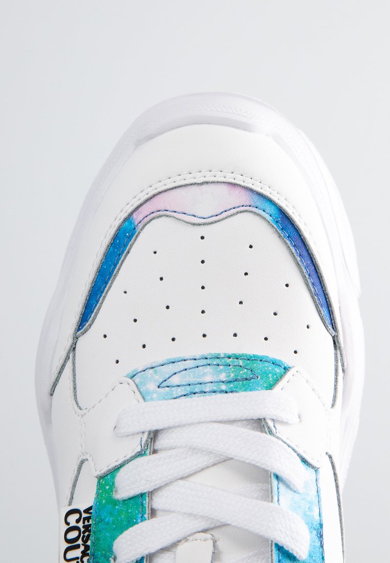 Zapatilla blanca con acentos abstractos y coloridos en azul y verde en la parte superior. Presenta detalles perforados, cordones planos y una puntera redondeada.