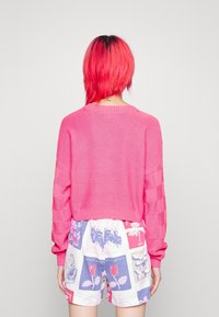 Kort rosa stickad tröja med texturerade rutmönstrade ärmar och en kortad nederkant, parat med mönstrade shorts med blommönster.