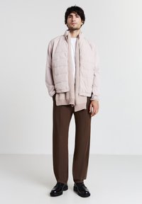 Helle rosa gepolsterte Jacke mit hohem Kragen, über einem beigen gerippten Pullover getragen, kombiniert mit braunen Hosen und schwarzen Schuhen.