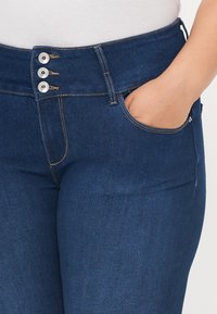 Tmavě modré džíny z denimu s vysokým pasem, opatřené třemi kovovými knoflíky nahoře, kontrastním šitím a detaily předních kapes.