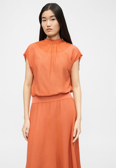 Jeune femme aux longs cheveux foncés portant une robe orange sans manches avec un col haut froncé et une taille cintrée.