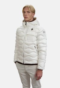 Giacca a vento bianca con cappuccio, caratterizzata da un design a righe orizzontali, chiusura con zip, tasche frontali e un logo sulla spalla sinistra.