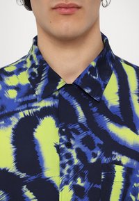 Close-up van een persoon die een overhemd draagt met een kraag, bedrukt met een abstract tie-dye patroon in blauw, zwart en neon geel, en een borstzak.