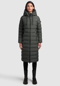 Manteau matelassé long et vert foncé avec une capuche, doté d'une fermeture éclair complète, d'un design à cloisons horizontales et de poches latérales. Porté avec des leggings noirs et des bottes.