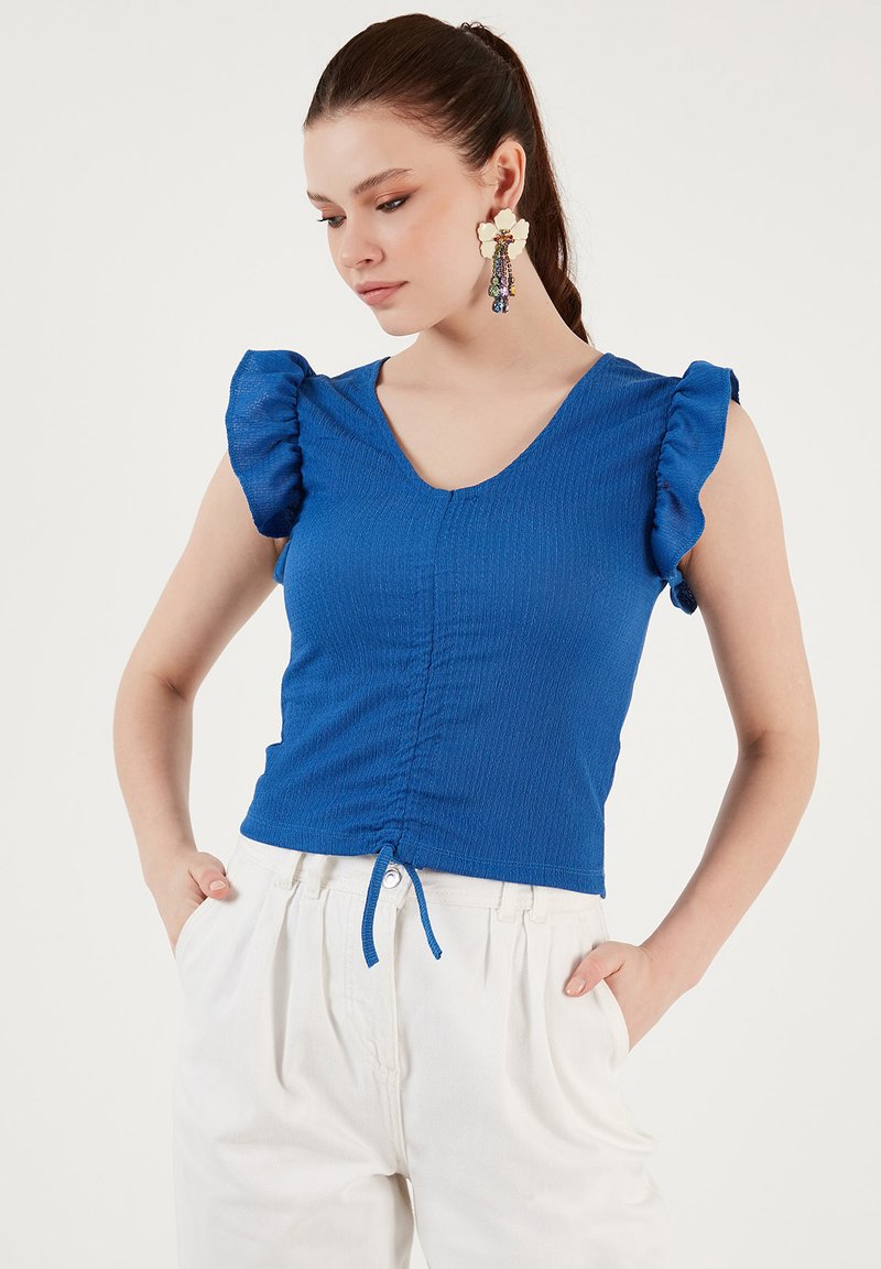Top bluetto con texture, maniche corte a volant, scollo a V e dettaglio arricciato frontale. Indossato con pantaloni bianchi a vita alta.