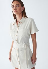 Beżowa sukienka koszulowa z krótkimi rękawami, dwiema kieszeniami na piersi, guzikami z przodu i paskiem w talii. Gładka tekstura i dopasowany krój.