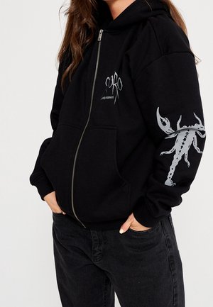 Femme portant un sweat à capuche noir zippé avec un motif de scorpion blanc sur la manche gauche et un petit logo sur la poitrine, mains dans les poches avant.
