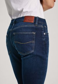 Mörkblå denimjeans med en brun läderpatch på midjan där det står "Pepe Jeans" präglat; klassisk femfickdesign med gyllene stygn.