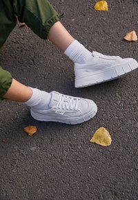 White sneakers with a thick sole, textured leather, and lace-up design, paired with white crew socks, resting on a dark asphalt surface.
