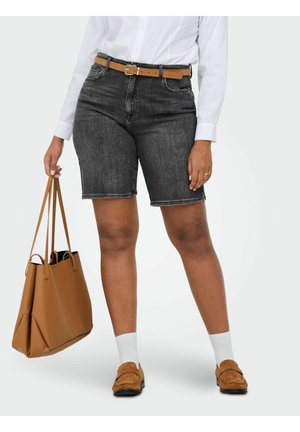 Femme portant un short en jean noir avec ceinture marron clair, chemise blanche à manches longues, chaussettes blanches, mocassins marron clair, et tenant un grand sac à main marron clair.