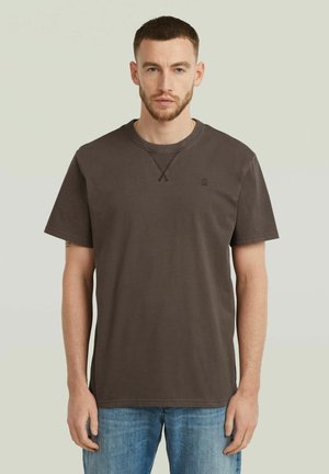 Jonge man met kort haar en baard, gekleed in een effen donkerbruin T-shirt met korte mouwen en lichtblauwe spijkerbroek, staand tegen een neutrale achtergrond.