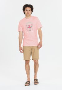 Vaaleanpunainen lyhythihainen paita, jossa on värikäs flamingokuvio, yhdistettynä vaaleisiin lyhyisiin housuihin ja ruskeisiin sandaaleihin.