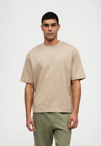 Beige short-sleeve t-shirt with a round neck, made of smooth fabric, featuring a relaxed fit and minimal stitching details. Paired with green pants.