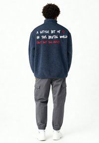 Veste en polaire bleu marine avec un col montant, comportant un texte rouge et blanc au dos. Associée à un pantalon cargo gris et des baskets blanches.