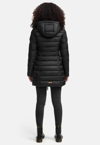 Doudoune noire avec capuche, présentant un design matelassé horizontal, des poignets élastiques et un ourlet à cordon de serrage. Portée sur un jean skinny noir et des bottes.