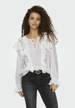 Weißes Blusenshirt mit Rüschendetails, Schnürung vorne und langen Ärmeln. Kombiniert mit schwarzen Jeans und einem Gürtel mit einer runden Schnalle.