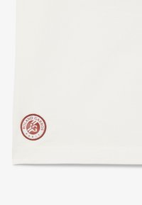 T-shirt in cotone bianco con un logo ricamato rosso rotondo che riporta la scritta "ROLAND GARROS" e un'icona sull'orlo. Texture liscia, vestibilità casual.