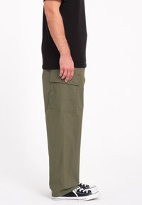 Pantalons cargo vert olive avec une coupe relaxe, dotés d'une poche latérale et d'une texture lisse. Associés à des baskets noires.