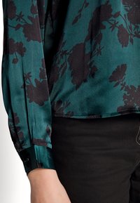 Długo rękawowa błękitna bluzka z czarnym kwiatowym wzorem, o gładkiej teksturze, z zapięciem na guziki przy mankietach i lekko zaokrąglonym dole. W połączeniu z czarnymi spodniami.